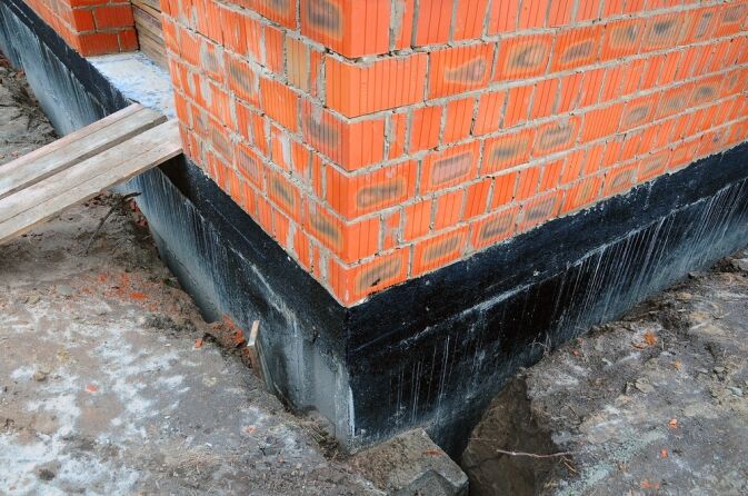 Brick wall corner of a building under construction, with black waterproofing applied to the concrete foundation. Dirt and wood planks are visible around the structure.
