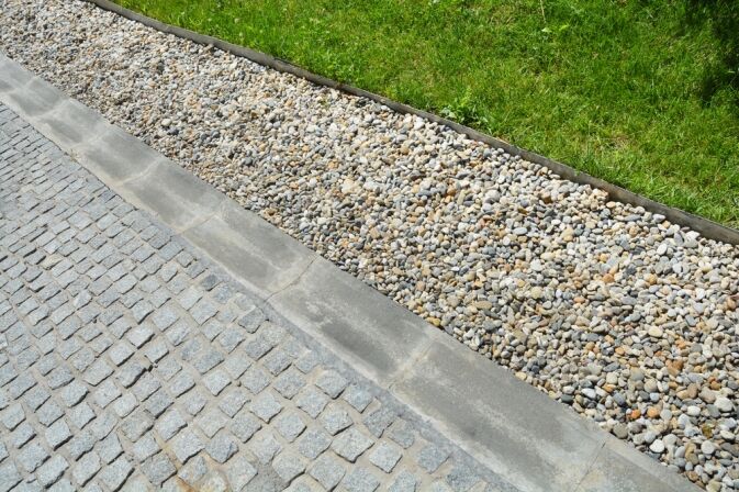 A section of pavement with gray cobblestones, a concrete curb, a strip of small rocks, and a grassy area.