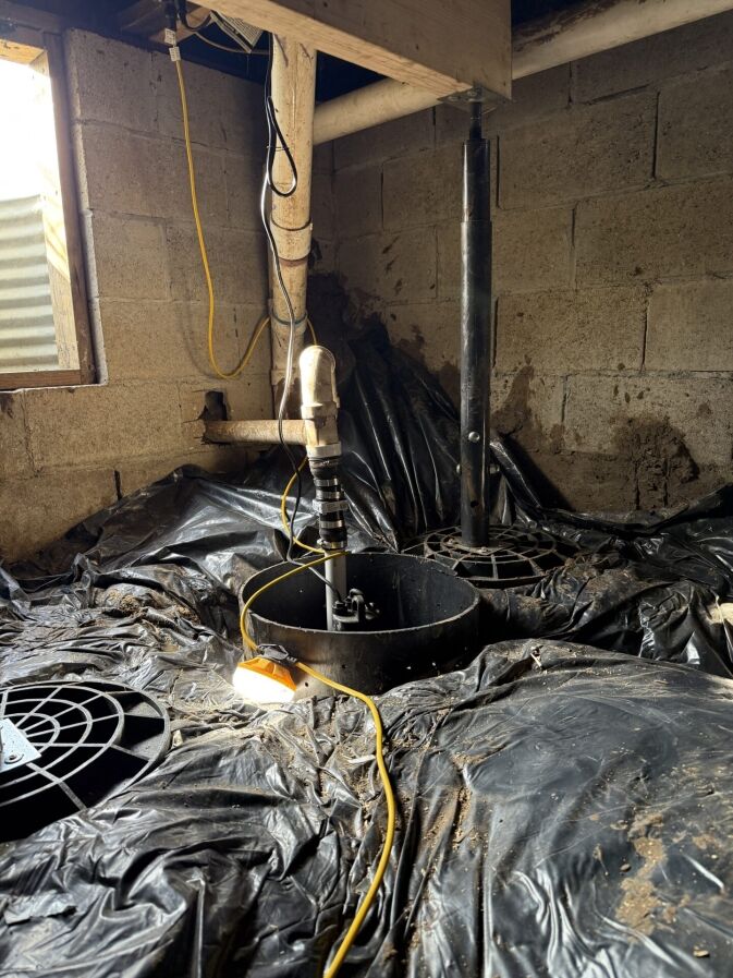A basement or crawlspace with exposed pipes, a sump pump system, plastic sheeting on the floor, and electrical cords running along the wall and ground.
