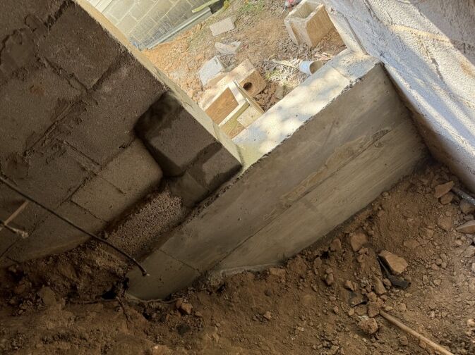 A partially constructed concrete wall with exposed soil and scattered construction materials is shown in a sunlit outdoor setting.