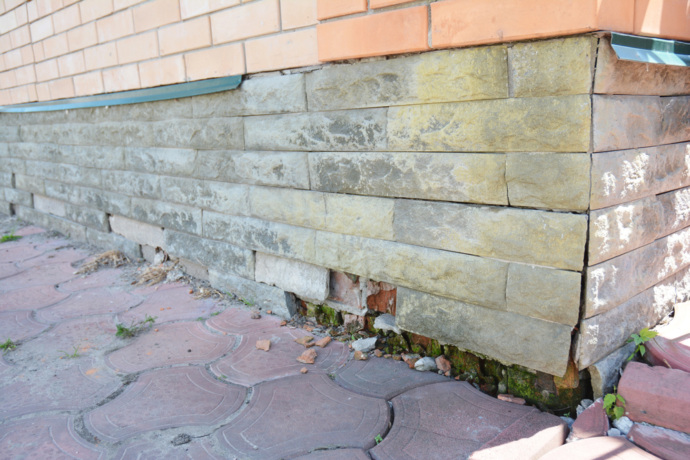A close-up of a building’s foundation showing cracked and damaged stonework with visible gaps and crumbling bricks at the base.