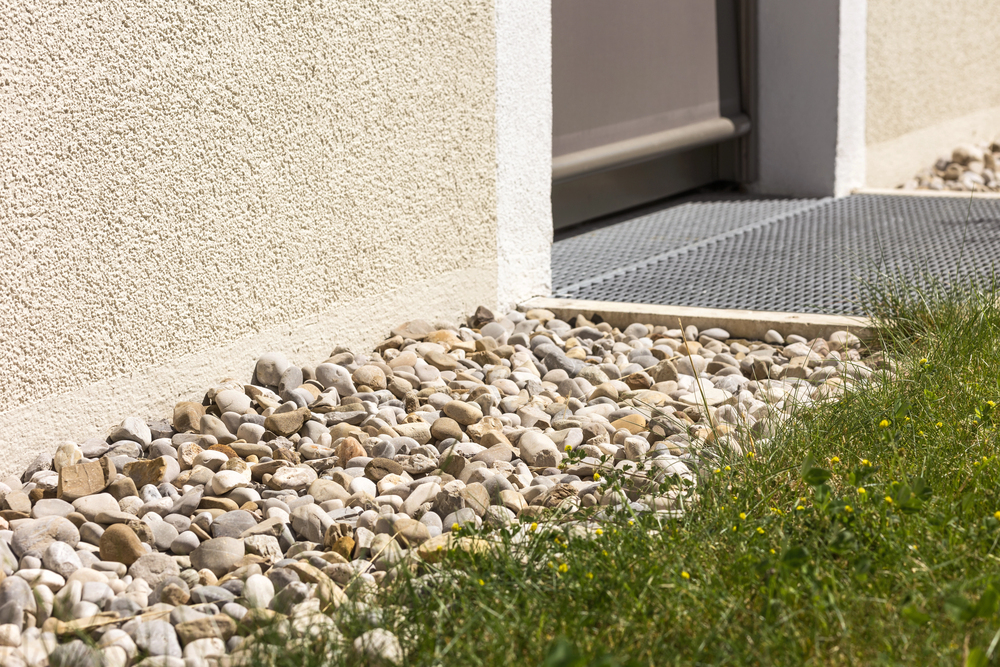 Close-up of a building’s exterior showing a gravel border next to a textured wall, a metal grate by a door, and green grass with small yellow flowers.