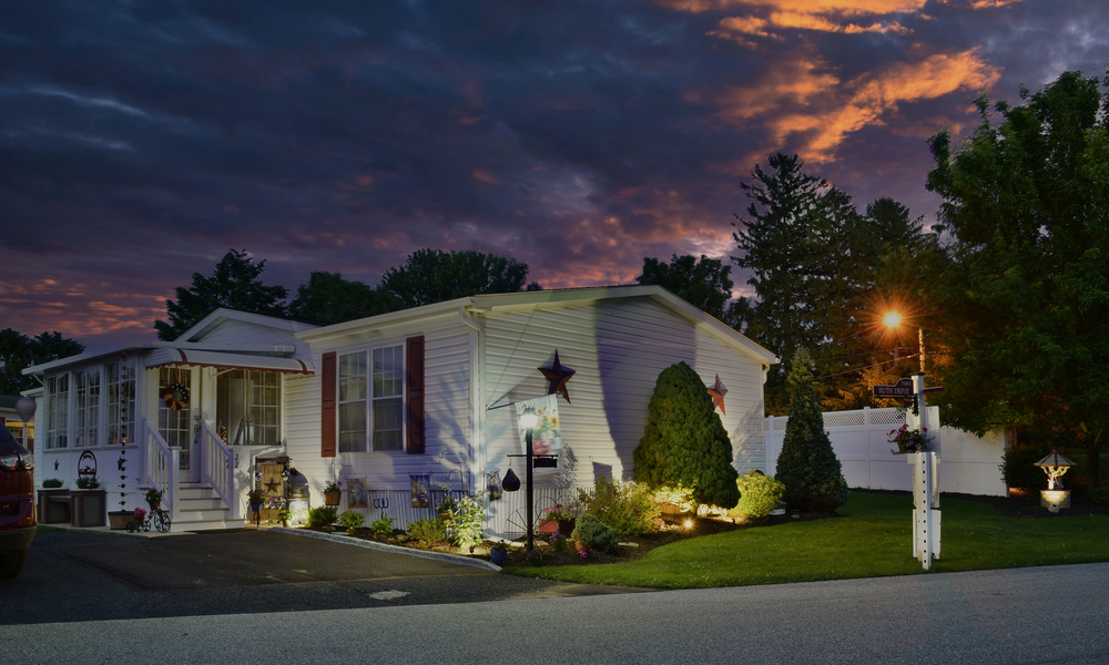 A white single-story house with a small garden and outdoor lights is pictured at dusk under a dramatic cloudy sky with orange and purple hues.